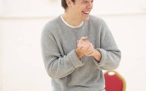 Jasper Talbot wears a grey sweater and blue jeans. He stands with his hands together and laughs, looking at something off camera.