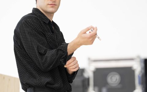 Arty Froushan wears black jeans and a black shirt. He stands, holding a tightly rolled note.