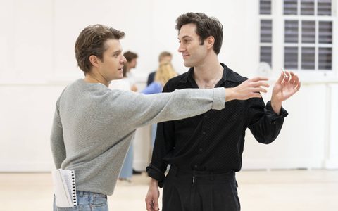 Arty Froushan wears black jeans and a black shirt. He stands facing Jasper Talbot, holding a tightly rolled note out of Jasper’s reach, who wears a grey jumper and blue jeans.