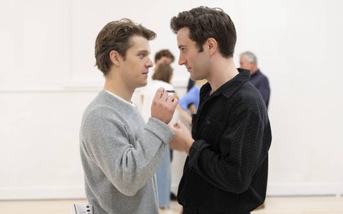 Jasper talbot wears a grey jumper and stands closely facing Arty Froushan, who wears a black shirt. Jasper holds a tightly rolled note.
