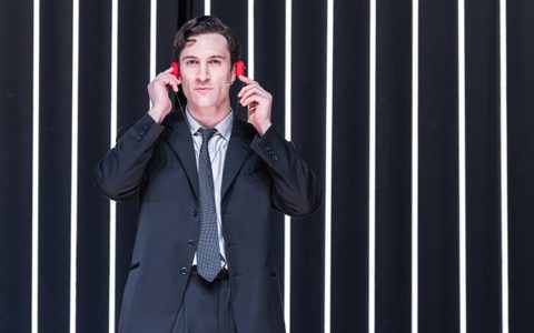 Arty Froushan wears a black suit with a light shirt and dark tie. He looks directly at the camera, wearing red headphones, to which his hands are raised.