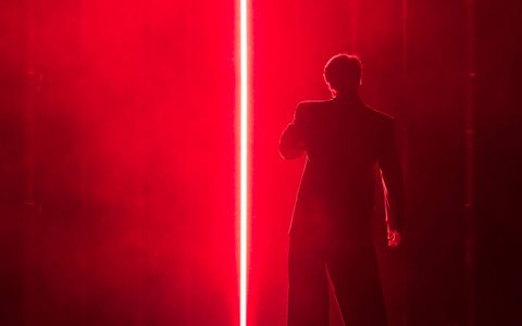 Arty Froushan wears a suit and faces away from the camera. He is bathed in red right, with a strip of white light on his left.