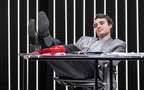 Oli Higginson wears a grey suit. He leans back with his feet up on a desk behind a red phone. He has a satisfied expression.