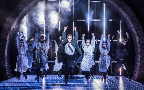 Arty Froushan leads the American Psycho company in choreography on stage. They each wear sunglasses and grey wraps over their suits, except Arty.