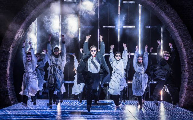 Arty Froushan leads the American Psycho company in choreography on stage. They each wear sunglasses and grey wraps over their suits, except Arty.