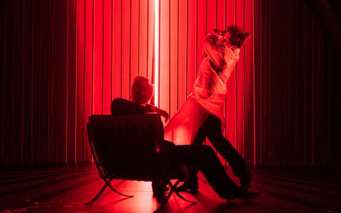 Arty Froushan wears a white plastic coat and swings an axed towards Daniel Bravo, who sits in a chair facing away from the camera. They are bathed in red light.