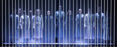 The American Psycho company stand in a row behind a curtain of light panels. They each wear sunglasses and grey wraps over their clothing.