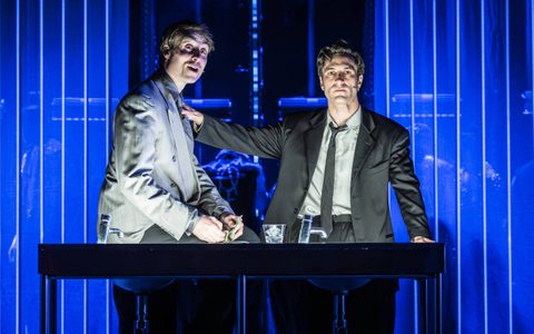 Arty Froushan stands behind a desk with his hand on Oli Higginson’s shoulder, he sits on the edge of the desk. They both wear suits. Oli has an excited expression.