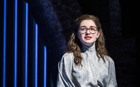 Anastasia Martin wears a metallic grey top and black pencil skirt, standing with her hands crossed in front of her. She looks off to the side with a worried expression.