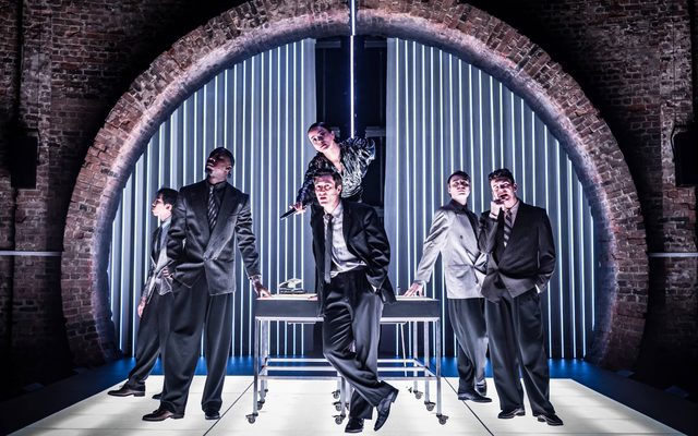 The American Psycho company stand around a desk on stage, illuminated by cool white-blue light.