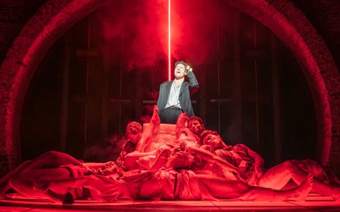 Arty Froushan wears a black suit with a light shirt and stands illuminated in white light. He holds a torch and sings, and in front of him other members of the company lay heaped around a chair, bathed in red light.