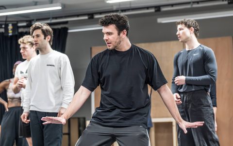 Jack Butterworth wears all black and poses with his hands out on either side of him. Alex James-Hatton, Arty Froushan, and Daniel Bravo stand in the background.