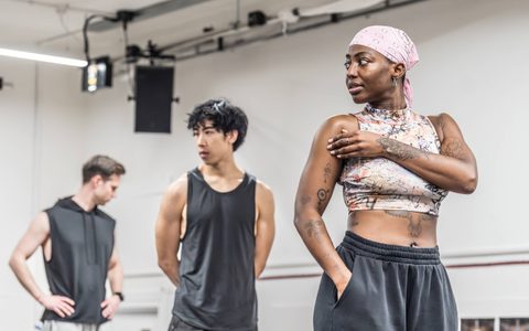 Liz Kamille wears a crop top and pink bandana, standing with one arm across her body and looking to the side. Zheng Xi Yong and Samuel J Weir stand in the background, blurred slightly.