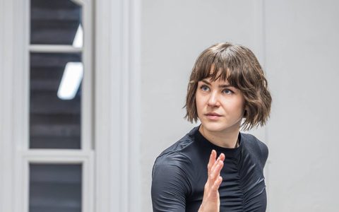Millie Mayhew stands in a running pose in a rehearsal room. She wears black.