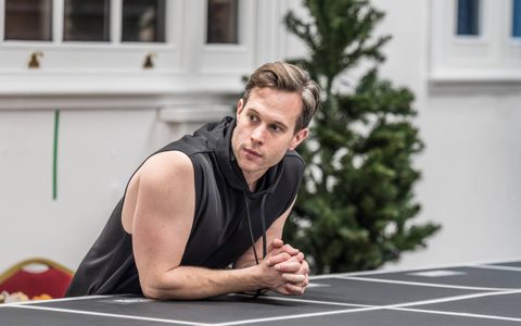 Samuel J Weir leans on a marked stage in a rehearsal room, looking to the side.