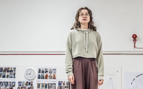 Anastasia Martin wears a green hoodie and brown trousers. She stands in a rehearsal room with a wary expression.