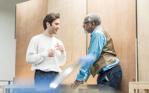 Arty Froushan wears a white top and stands talking to Joseph Mydell, who wears a blue shirt and brown waistcoat.