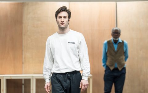 Arty Froushan wears a white top, standing with his hands by his side and looking straight ahead. Joseph Mydell stands blurred in the background.