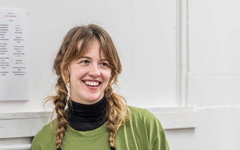 Associate Director Bethany West wears a green t-shirt. She is smiling, leaning against a wall.