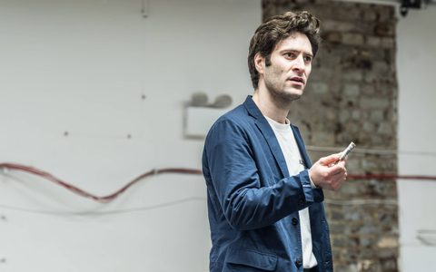 Arty Froushan wears a white top under a blue blazer. He looks towards someone off camera, holding up folded cash in front of him.