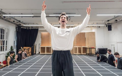 Arty Froushan wears a white top and stands at the front of a marked platform in a rehearsal room. He shouts, holding both hands up.