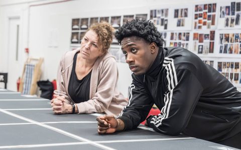 Posi Morakinyo and Kim Ismay lean forward, resting on a marked stage in a rehearsal room.