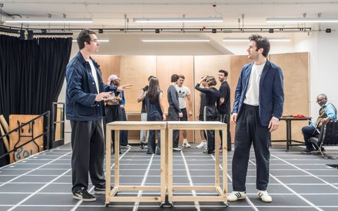 Oli Higginson and Arty Froushan each wear dark trousers, white tops and blue jackets. They face each other from across two tables in the centre of a rehearsal room. The American Psycho company stand in the background.