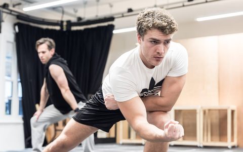 Daniel Bravo leans forward, resting on one knee with one fist raised in front of him, rehearsing choreography.