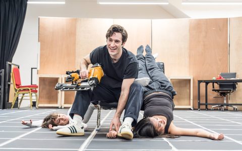 Arty Froushan sits holding a drill and leans forward, smiling. Millie Mayhew and Hannah Yun Chamberlain lie upside down behind him from the chair.