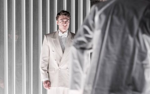 Daniel Bravo wears an off-white suit and white bow tie. He stands in front of a white panelled curtain, looking at some in front of, and turned away from, the camera.