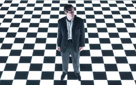 Arty Froushan wears a black suit with a light shirt and stands illuminated in white light. He looks to the side, photographed from above. The floor of the stage is a black and white checkerboard.