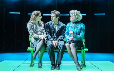 Arty Froushan wears a suit, sitting between Anastasia Martin and Kim Ismay on a bench. Each of the women wear smart jackets and skirts, and turn towards Arty. They are illuminated by blue and green light.