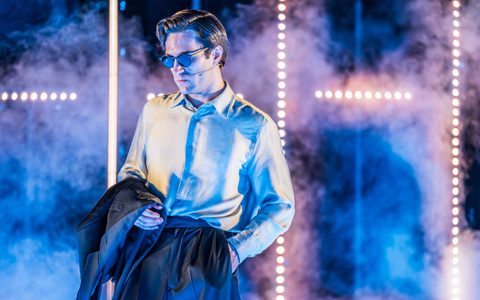 Arty Froushan wears black trousers with a light shirt, carrying a suit jacket over his arm. He wears sunglasses. The background is hazy and he is illuminated by blue light.