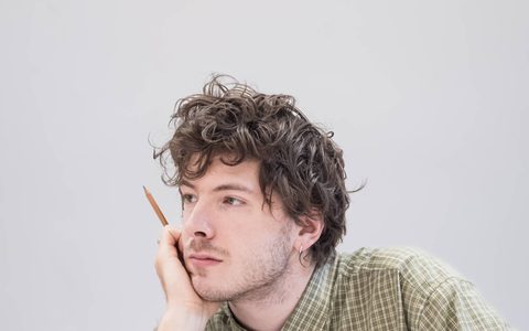 Sam Grabiner wears a green and white check shirt and sits with his chin resting in his hand.
