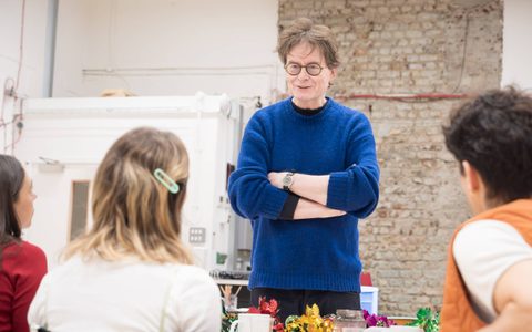James Macdonald wears a blue sweater. Smiling and with crossed arms, he stands in front of the Callie Cooke, Jacob Fortune-Lloyd, and Bel Powley, who have their backs to the camera.