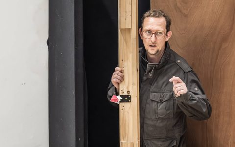 Leo Bill leans around a wooden beam and points forward.