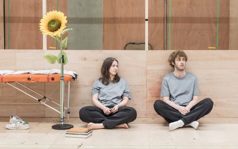Pearl Chanda and Jack Riddiford sit cross legged on a wooden floor. There is a large yellow sunflower next to them.