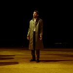 Josh O'Connor stands on a yellow floor, against a black background. His eyes are closed, and his left hand is partially splayed.