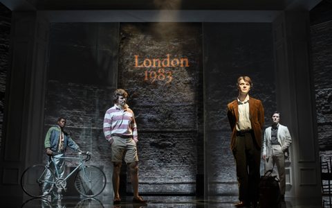Jasper Talbot wears a brown suit and stands on stage, looking ahead. Arty Froushan, Alistair Nwachukwu, and Leo Suter stand looking at him from behind. The text ‘London 1983’ is projected in orange on the wall.