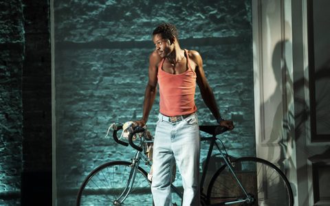 Alistair Nwachukwu wears an orange vest top and blue jeans. He leans back on a bicycle and looks to the side with a mischievous expression.