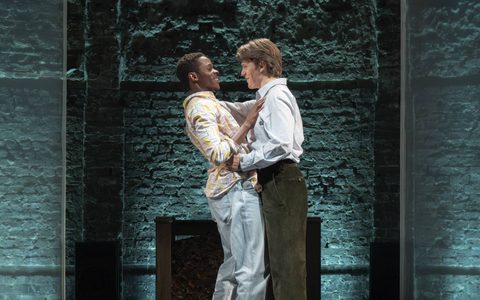 Alistair Nwachukwu and Jasper Talbot hold each other close, smiling at one another. Alistair wears a loose shirt, blue jeans and trainers. Jasper wears a formal light shirt, dark green trousers and brown loafers.