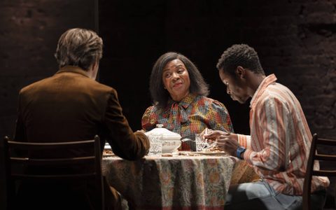 Doreene Blackstock wears a colourful printed top, and smiles across a dinner table at Jasper Talbot, who wears a brown blazer and face away from the camera. Alistair Nwachukwu sits between them, wearing a white and orange shirt.