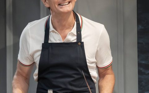 Charles Edwards wears a white polo shirt and dark apron. He smiles, standing behind a table to prepare drinks.