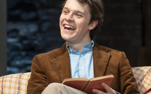 Jasper Talbot wears a blue shirt and brown corduroy blazer. He sits on a sofa, smiling and holding an open book in his lap.