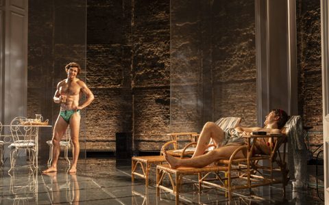 Leo Suter wears green Speedos and stands near an outdoor table on stage. He has one hand on his hip and looks towards Jasper Talbot, who smiles at him from a sun lounger.