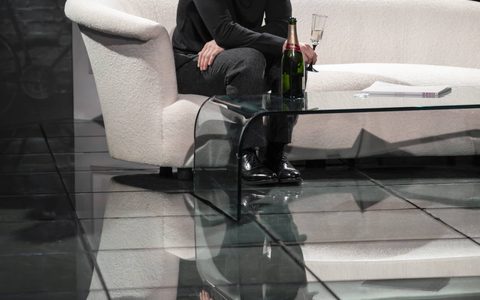 Arty Froushan wears all black and sits on a white sofa on stage, holding a champagne flute. He has a worried expression.