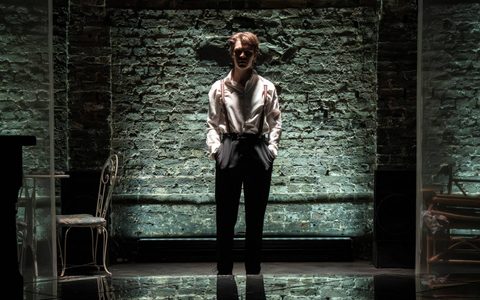 Jasper Talbot stands alone on stage. He has his hands in his pockets and is dimly lit.