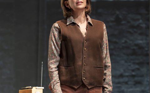 Claudia Harrison stands near a table on stage. She wears a brown waistcoat and skirt.