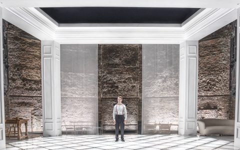 Jasper Talbot stands alone on a brightly lit stage. The set is a grand house, with four white pillars at each corner.