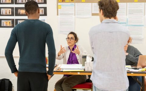 Sophie Dillon Moniram wears a purple top and light cardigan. She sits at a table, talking to Alistair Nwachukwu and Jasper Talbot, who have their backs to the camera.
